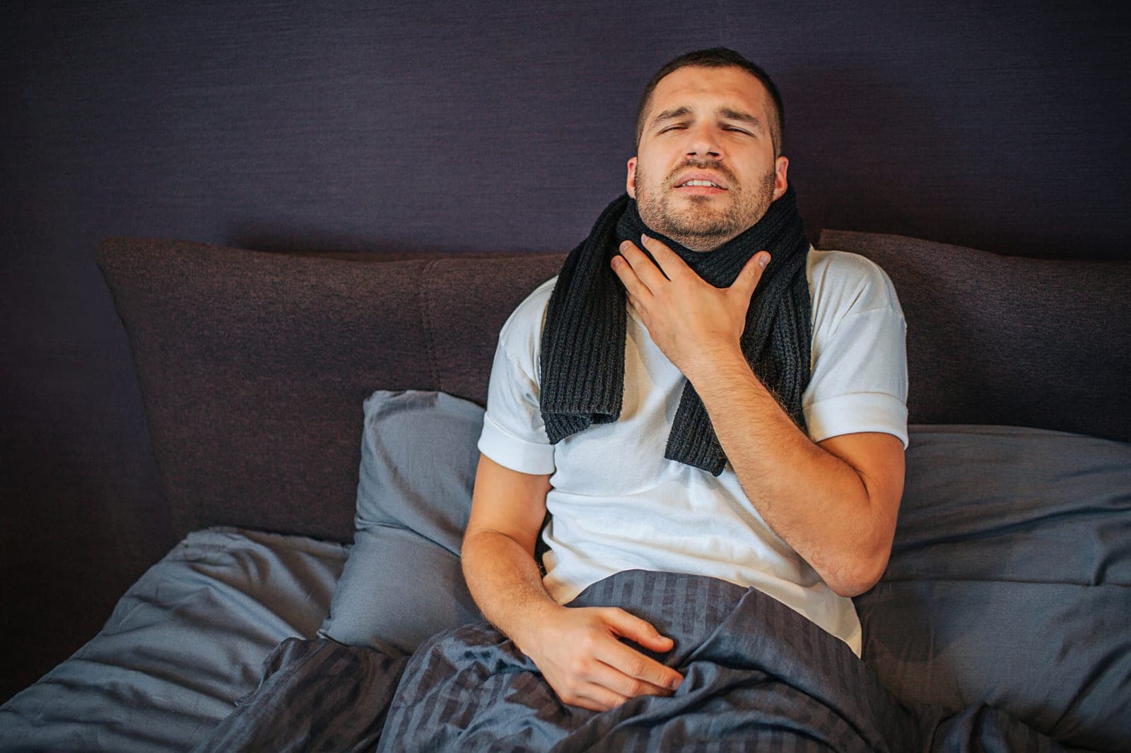 Homem na cama com cachecol, com a mão na garganta e expressão de dor