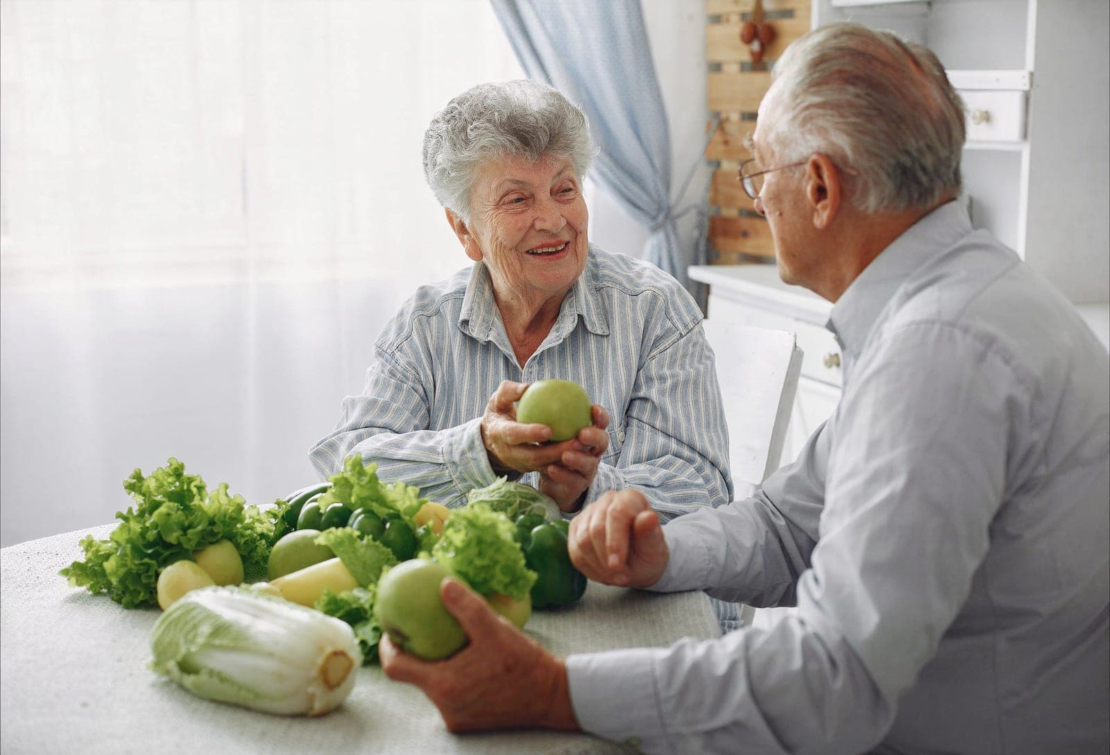 Casal de idosos sorrindo enquanto preparam uma refeição saudável com vegetais e frutas frescas